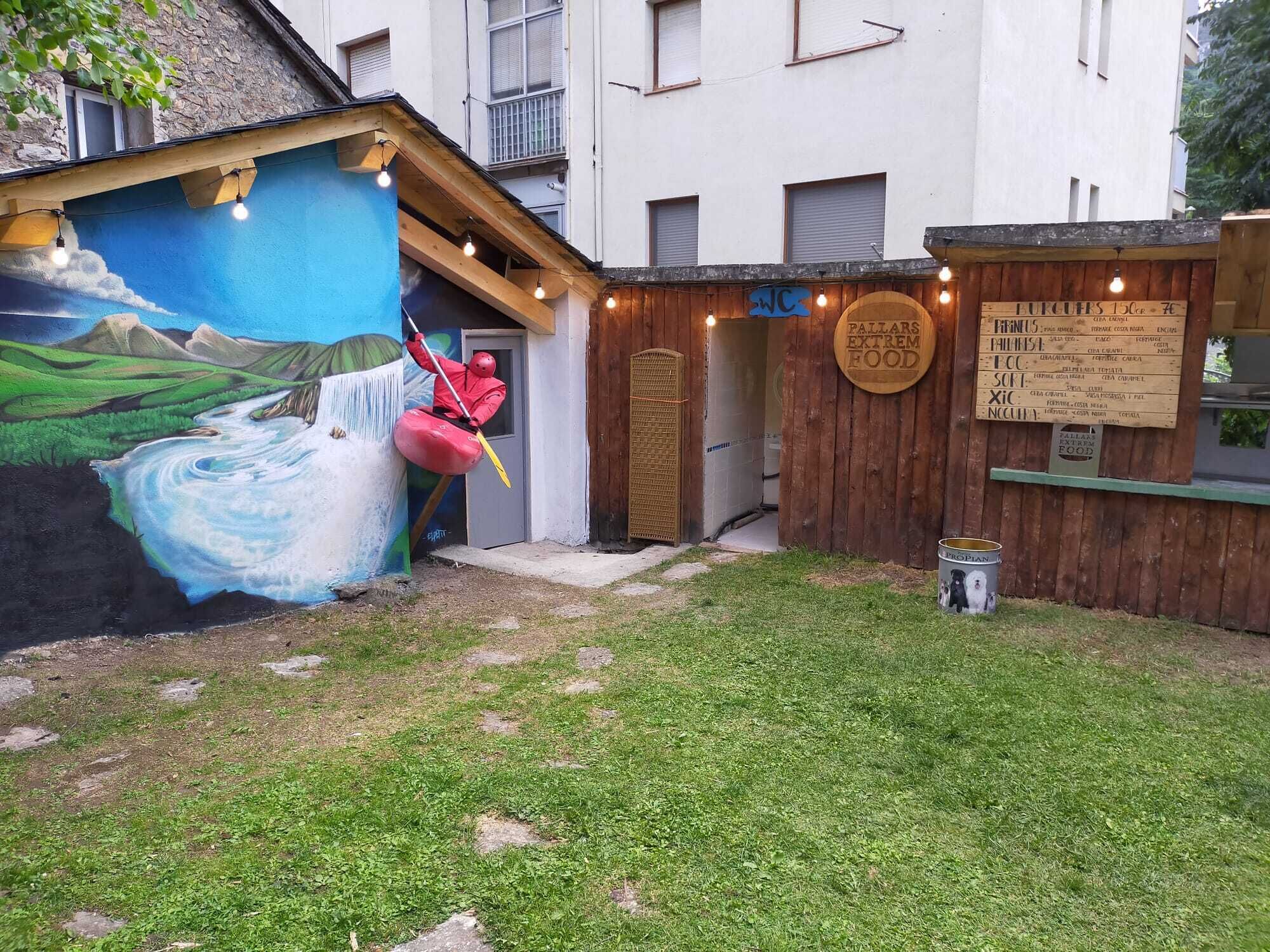 A rustic outdoor area with a mural of a river and mountains, a wooden structure labeled "Pallars Extrem Food", and a sign listing food items.