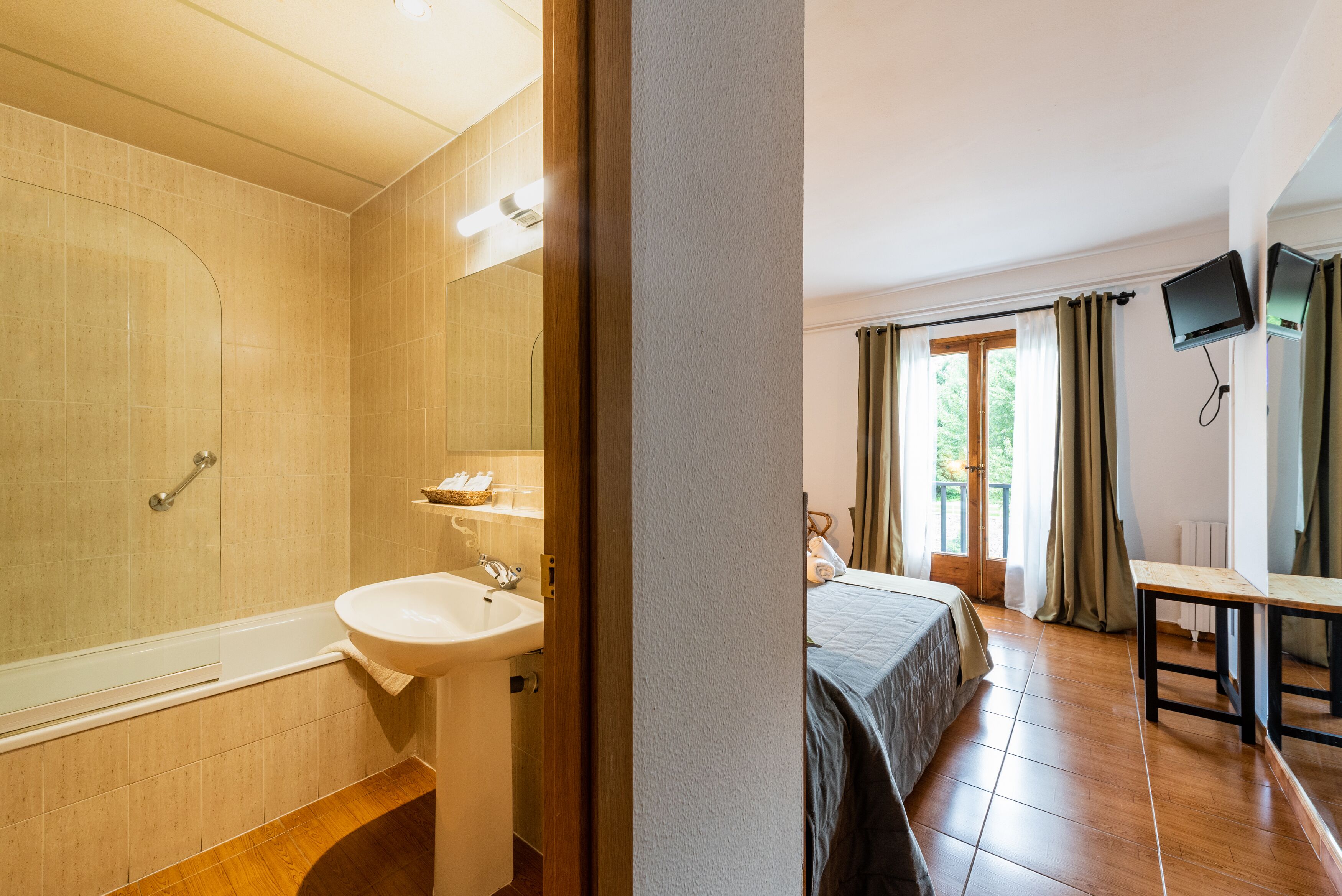A bathroom with a bathtub, sink, and mirror. There is a bed with a window and curtains in the adjacent room.