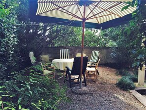 Een terras buiten met een ronde tafel en gestreepte stoelen, omgeven door weelderige begroeiing en een grindpad.