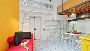 A compact living space with a red sofa, a dining table with chairs, a kitchen area, and a 'WELCOME' sign on the wall.