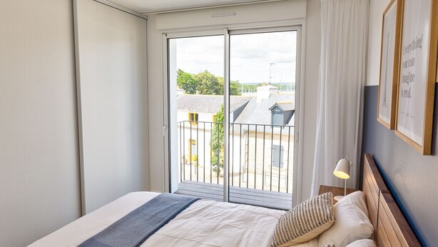 A hotel room with a large sliding glass door leading to a balcony, a bed with a blue throw blanket, and a nightstand with a lamp. - Port Ste-Marine: L'Escale - Balconies & Small view (Combrit)