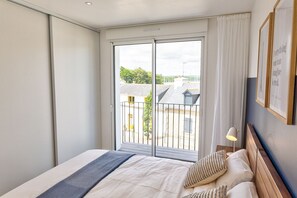 Una camera d'albergo con un'ampia porta-finestra che si apre su un balcone, un letto con copriletto blu e un comodino con una lampada.