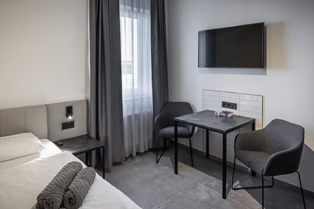 A hotel room with a bed, a desk, two chairs, a flat-screen TV, and a window with curtains.