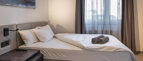 A neatly made bed with white linens, a small bedside table with a remote control, and a painting on the wall.