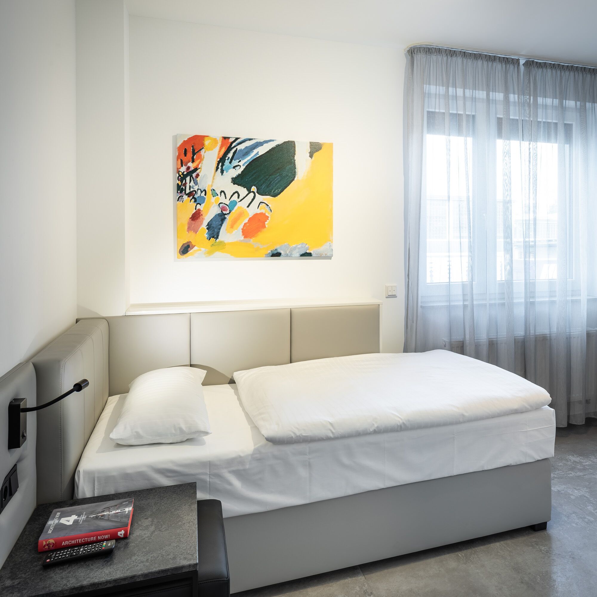 Una habitación de hotel moderna con una cama, una mesita de noche con un libro y un control remoto, un cuadro en la pared y una ventana con cortinas traslúcidas.