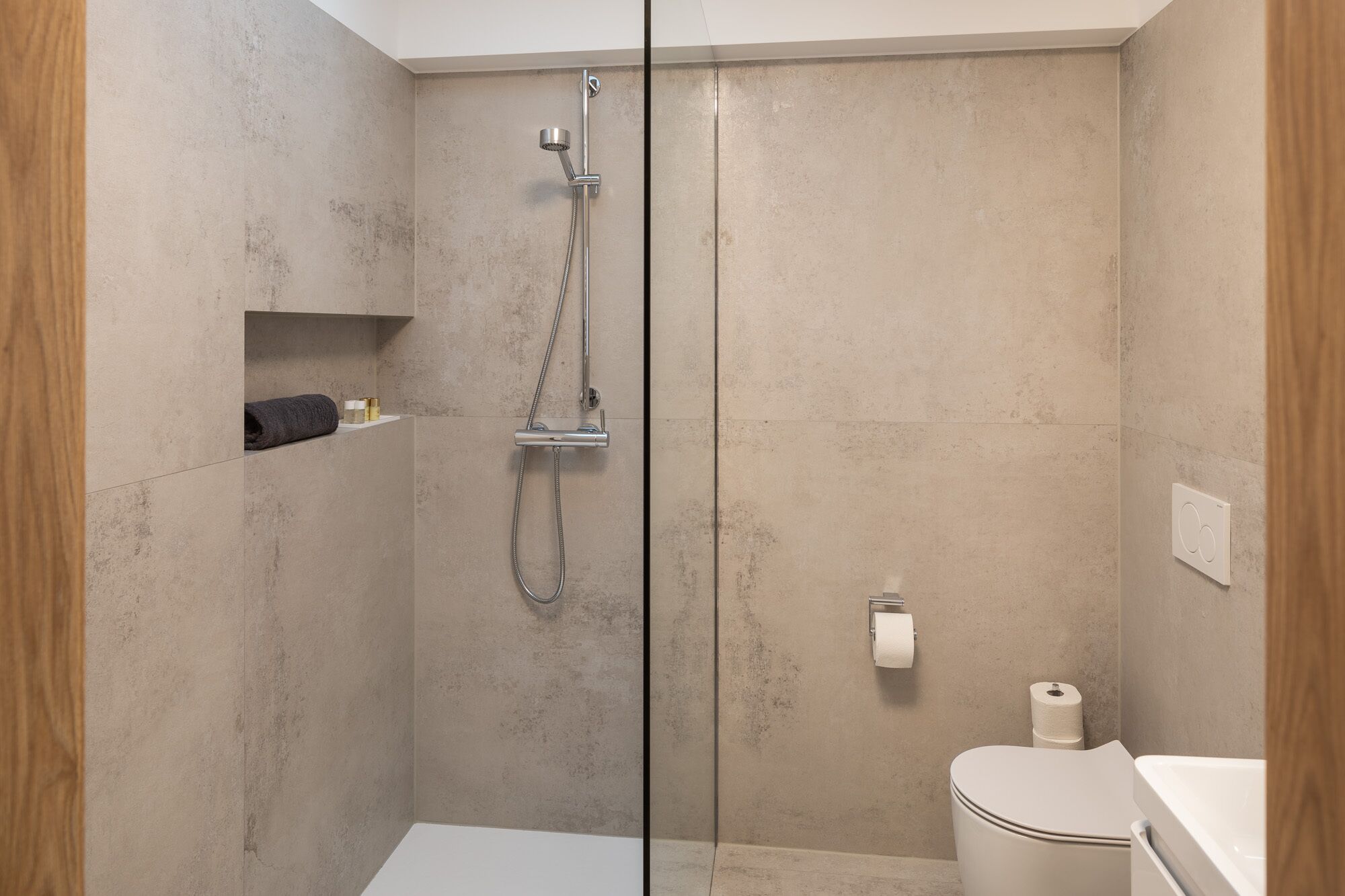A modern bathroom with a glass shower enclosure, a toilet, and a small shelf with toiletries.