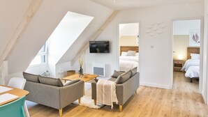 Una habitación moderna estilo loft, con techo alto, televisor de pantalla plana en la pared, un conjunto de sofás, una mesa de centro de madera y una mesita auxiliar.