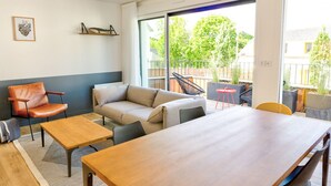 A modern living room with a sofa, armchair, and coffee table. There is a balcony with outdoor seating and a view of greenery. - Les Docks (Combrit)