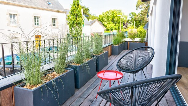 A balcony with black chairs, a red table, and planters with green plants. - Les Docks (Combrit)