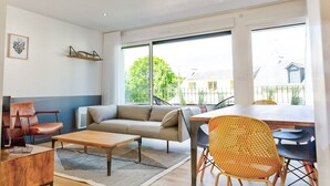 A modern living room with a sofa, coffee table, and chairs, large windows with a view of greenery and buildings, and a dining area with a table and chairs. - Les Docks (Combrit)