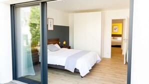 A modern bedroom with a large sliding glass door leading to a balcony, a bed with white linens, a bedside table with a lamp, and a framed picture on the wall. - Les Docks (Combrit)