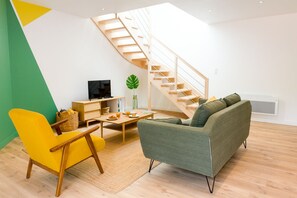 A modern living room with a wooden staircase, a yellow armchair, a green sofa, and a wooden coffee table.