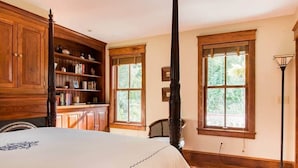 A bedroom with a large bed, wooden bookshelves, a tall black column, and large windows.