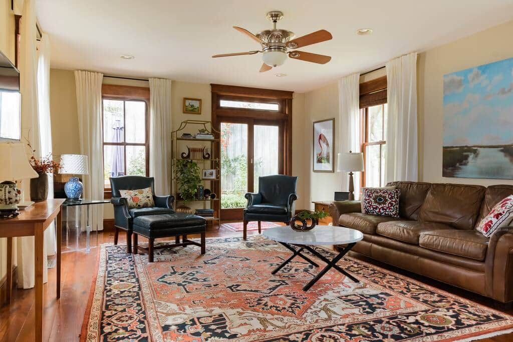 A living room with a ceiling fan, a brown leather sofa, a coffee table, two armchairs, a patterned rug, and a bookshelf.
