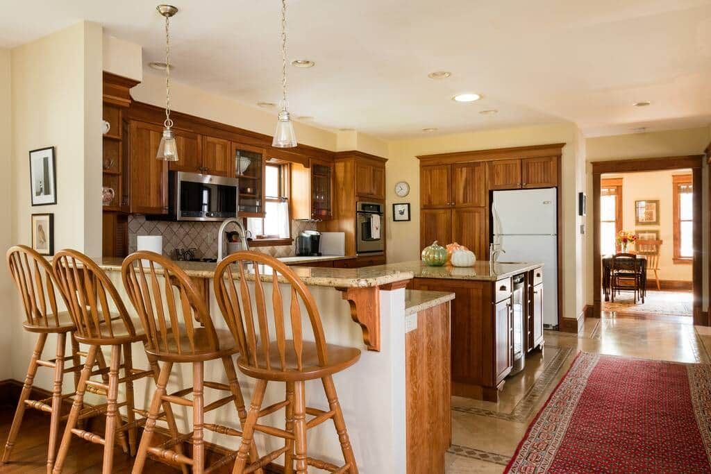 A kitchen with wooden cabinets, a central island with bar stools, stainless steel appliances, and a red patterned rug.