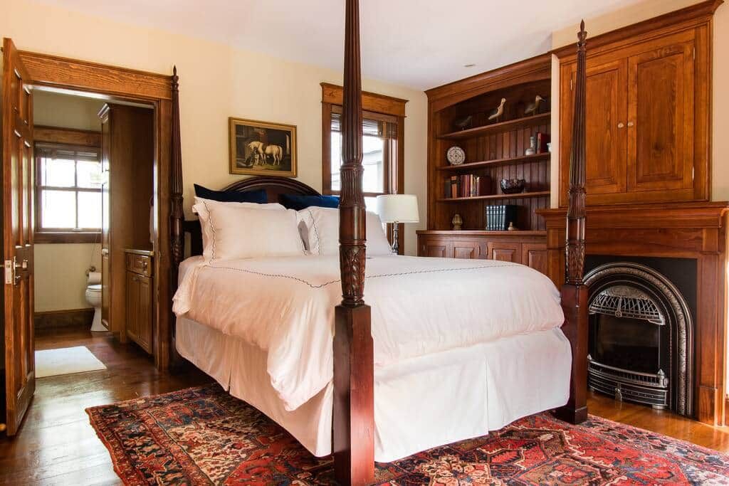 A four-poster bed with a white bedspread, a red and black patterned rug, a wooden bookshelf with books and decorative items, and a fireplace.