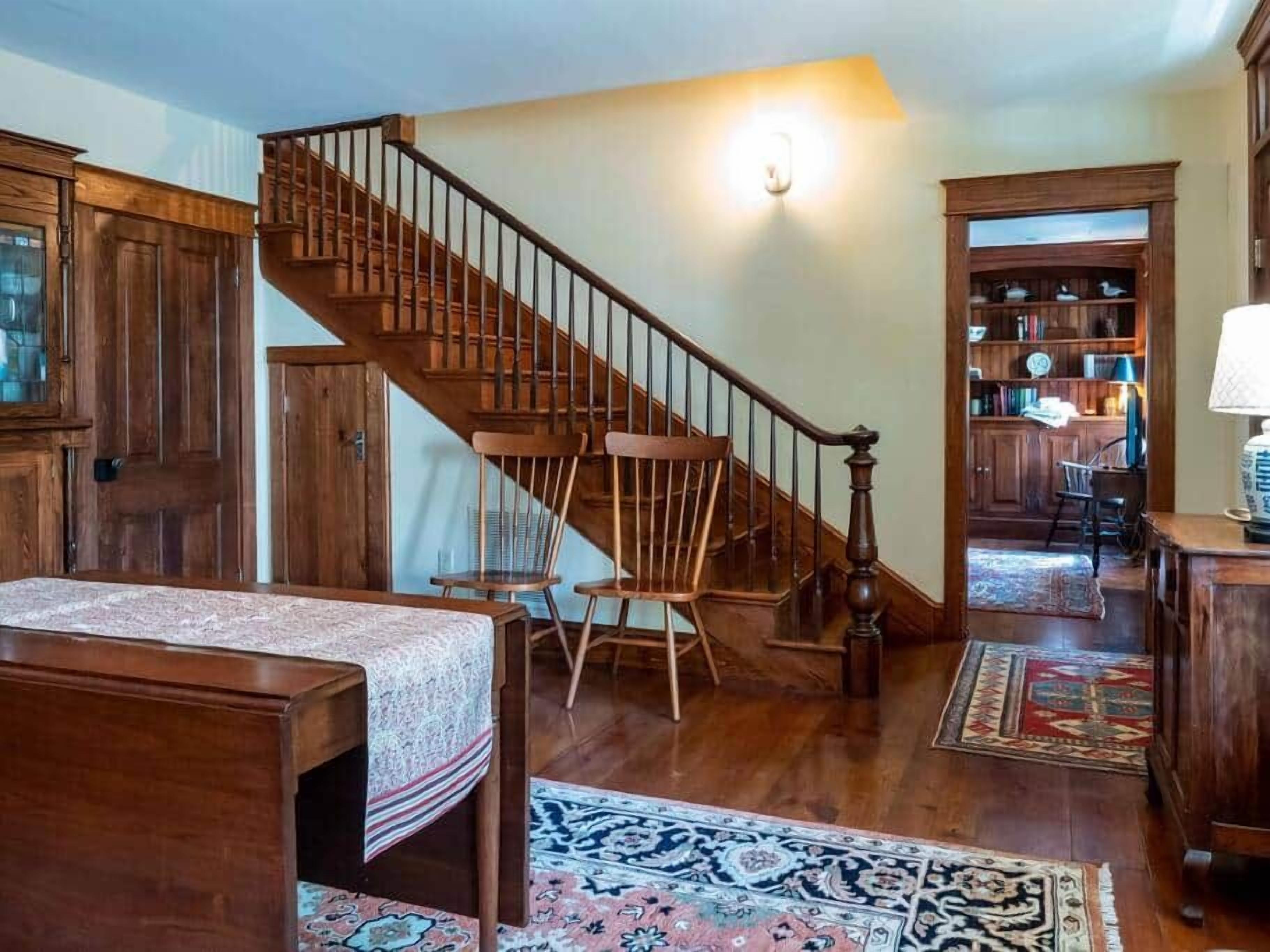 A wooden staircase leading to a second floor, a large wooden bookcase, and a patterned rug on the floor.