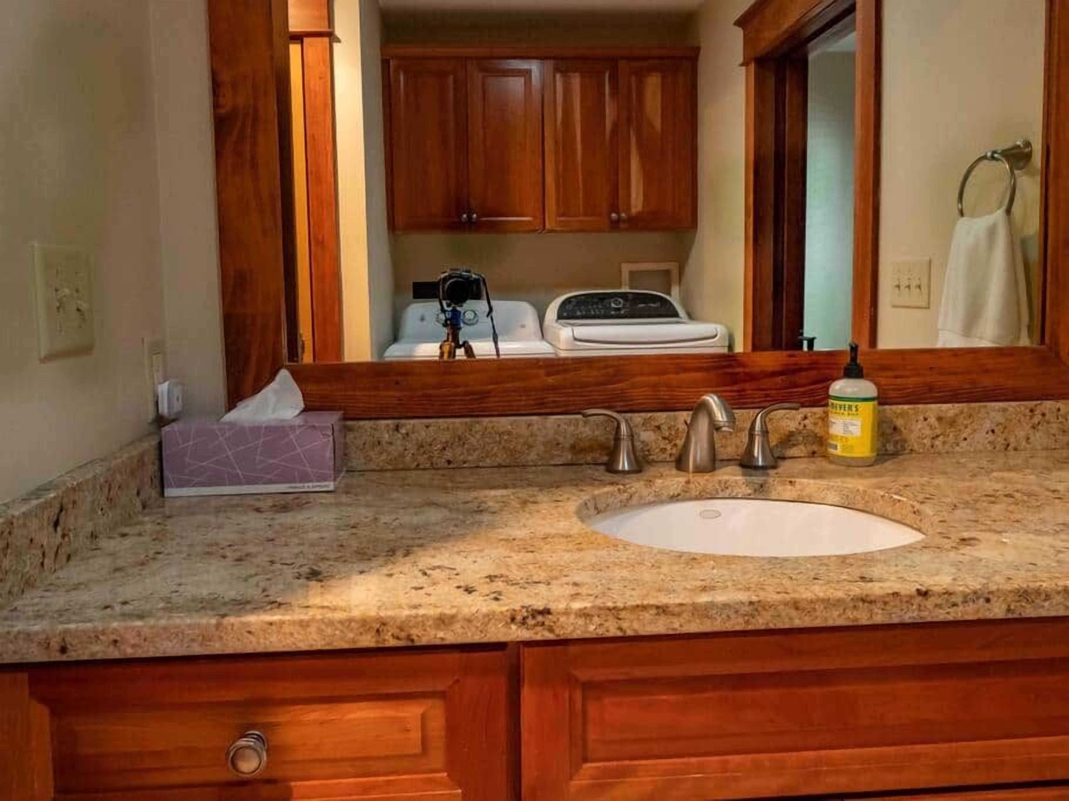 Une salle de bain dotée d’un comptoir en granit, d’un lavabo blanc, d’armoires en bois et d’un miroir qui reflète la pièce.