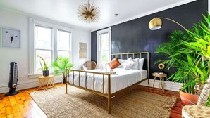 Ein Schlafzimmer mit einem Bett, einem großen Fenster, einem Standventilator und einer Topfpflanze.