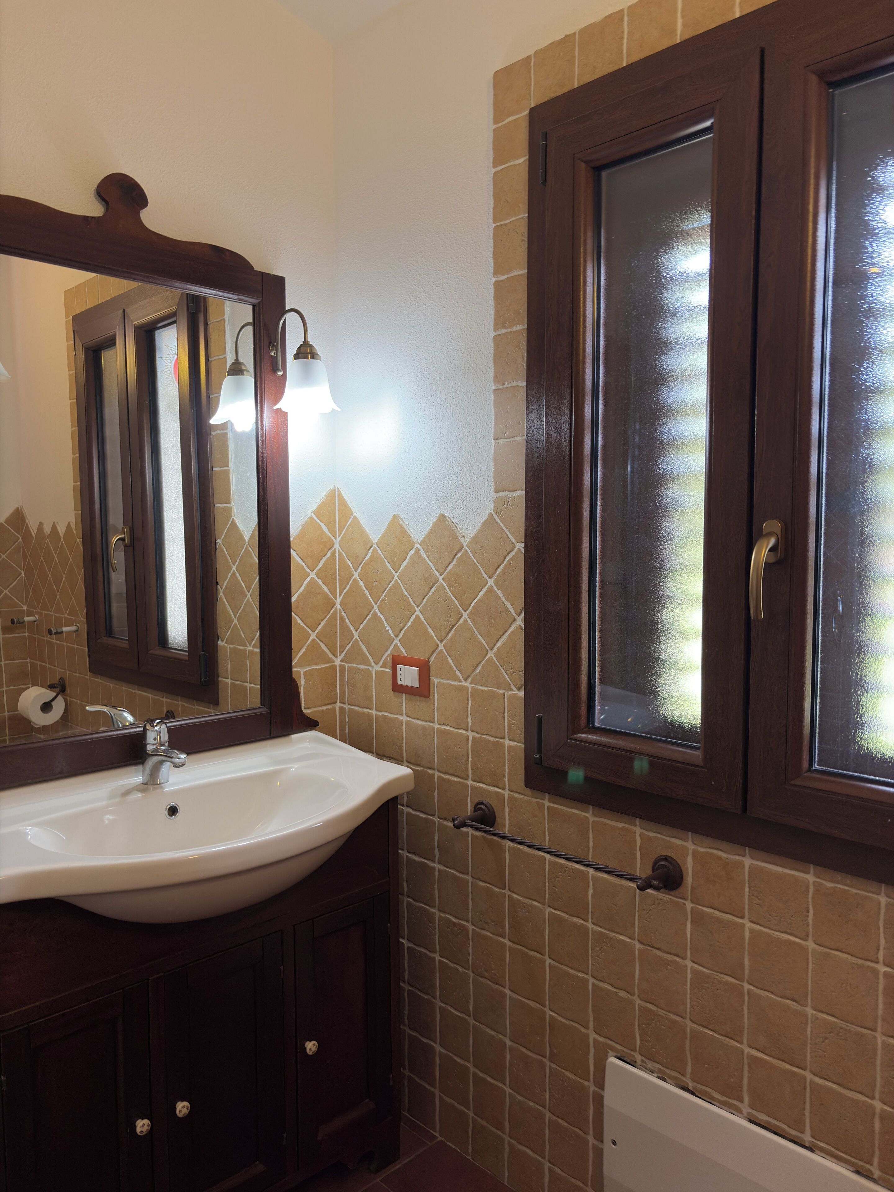 Une salle de bain avec un lavabo blanc, des meubles en bois foncé, un miroir et une fenêtre à vitres dépolies.