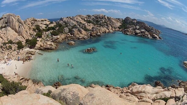 Une zone côtière rocheuse avec une eau turquoise cristalline, une plage de sable et des gens qui se baignent.