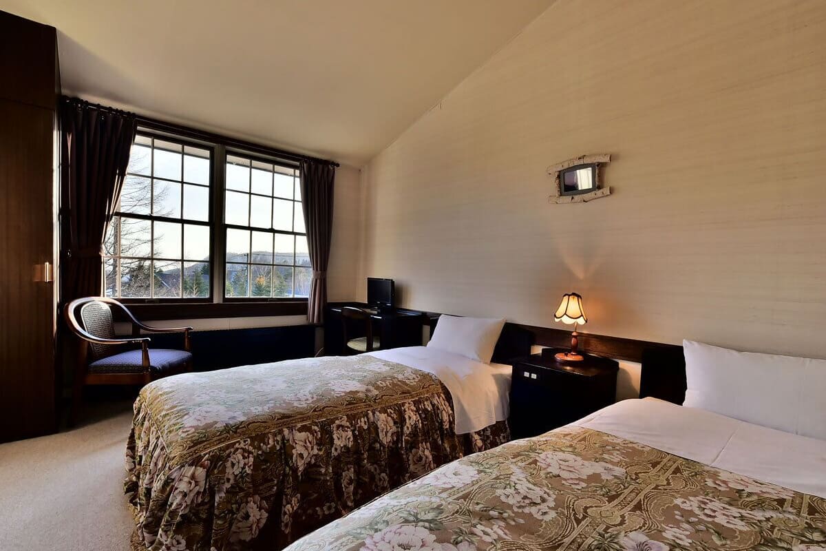 Ein Hotelzimmer mit zwei Betten, einem Schreibtisch, einem Stuhl, einer Lampe und einem Fenster mit Vorhängen.