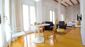Un salón luminoso con una mesa blanca, sillas y un sofá. Hay ventanas que dejan entrar luz natural, un techo de madera y una pared con un estampado.