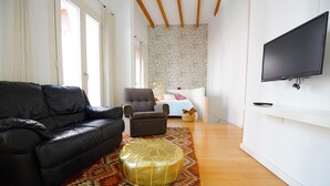 Una sala de estar con un sofá de cuero negro, un taburete dorado, un sillón marrón, una mesita de noche blanca y un televisor montado en la pared.