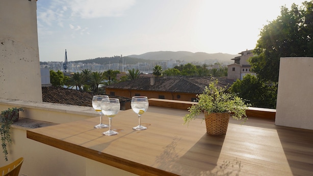 Un balcón con dos copas de vino blanco, una planta en maceta y una vista de un paisaje urbano con edificios y montañas a lo lejos.