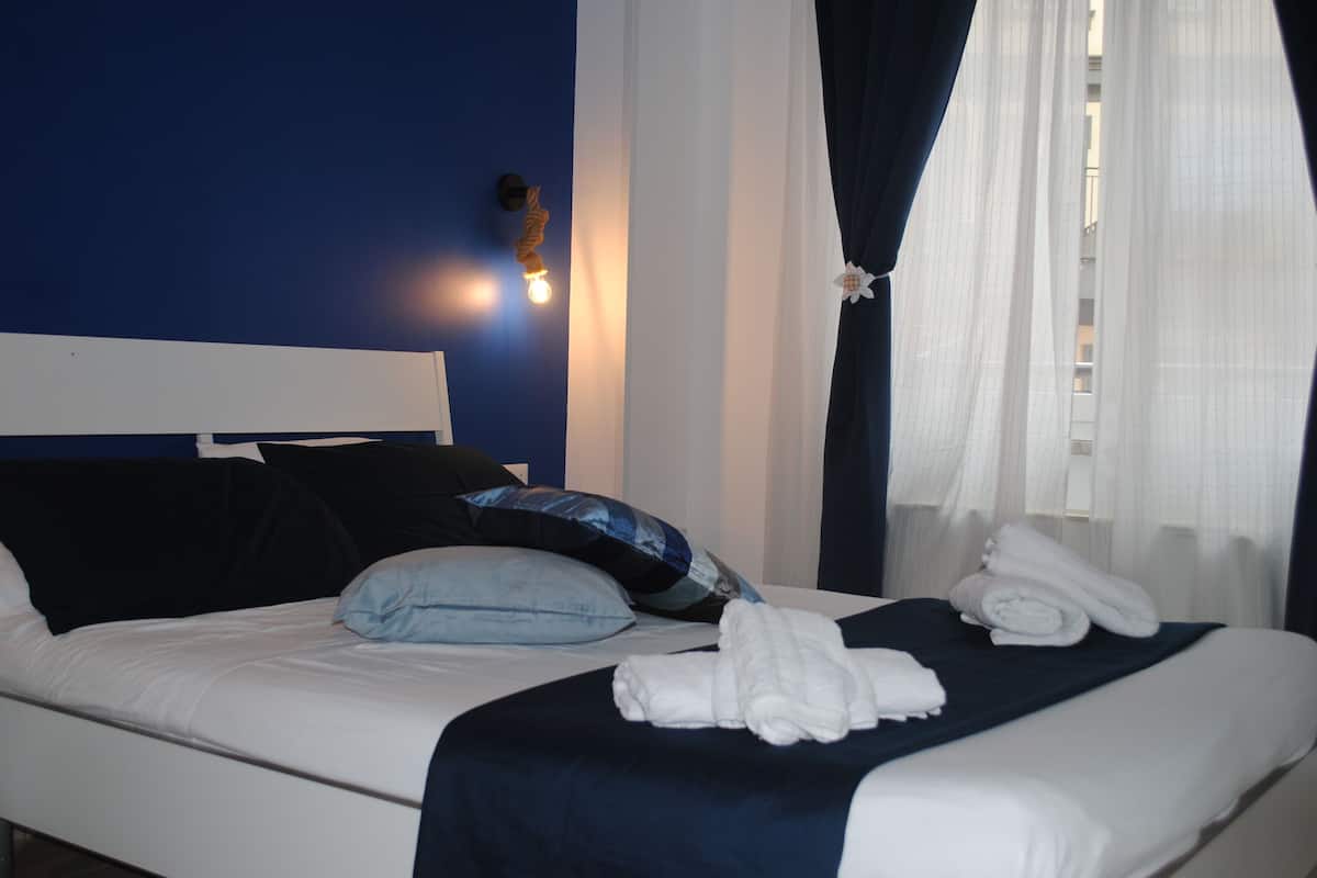 Una cama bien hecha con sábanas blancas, cabecera azul oscuro y almohada azul. Hay dos toallas blancas dobladas sobre la cama.