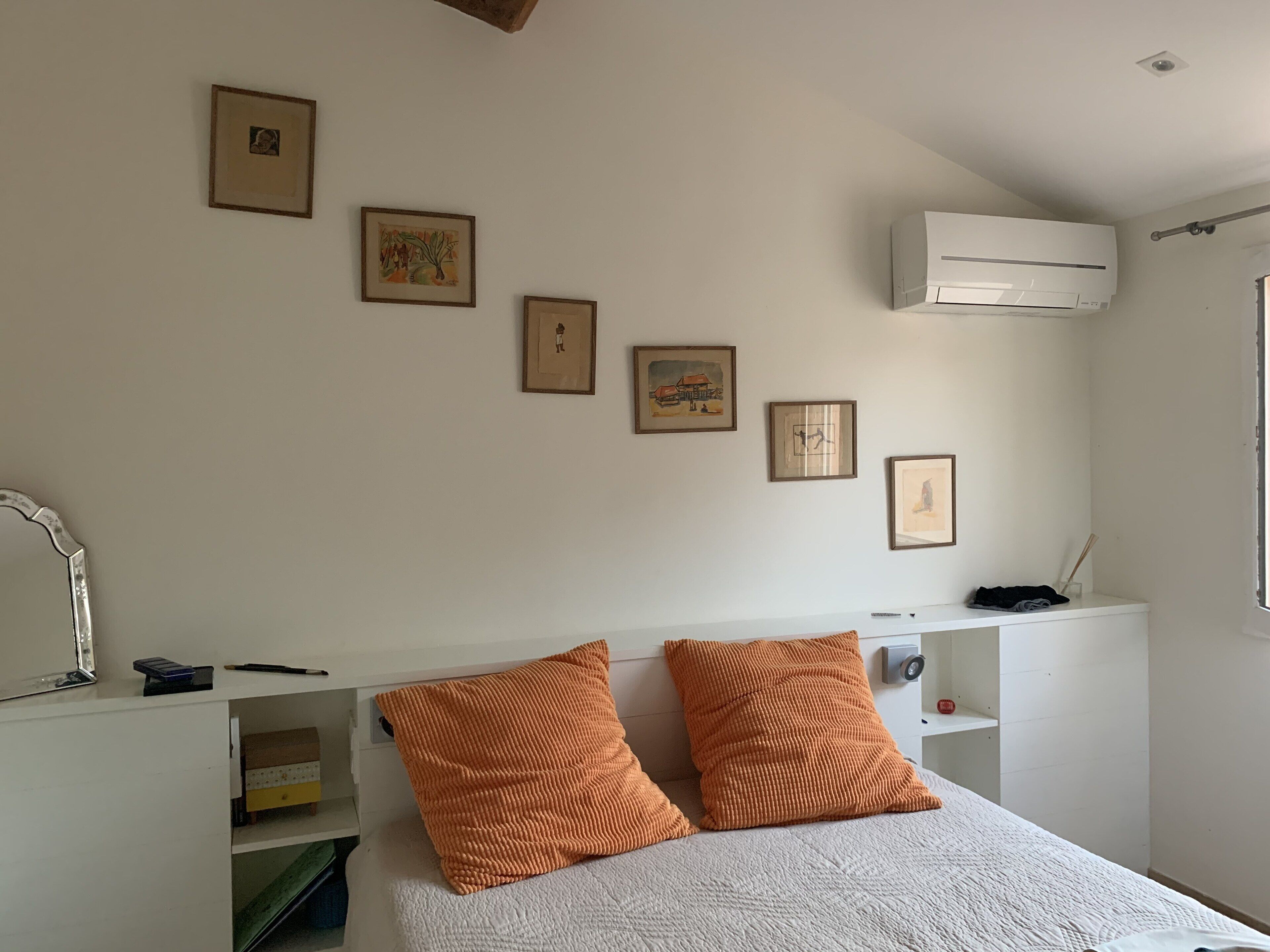 Ein Schlafzimmer mit einem Bett, zwei orangefarbenen Kissen, einem weißen Kopfteil, einem weißen Regal mit einer Fernbedienung und mehreren gerahmten Bildern an der Wand.