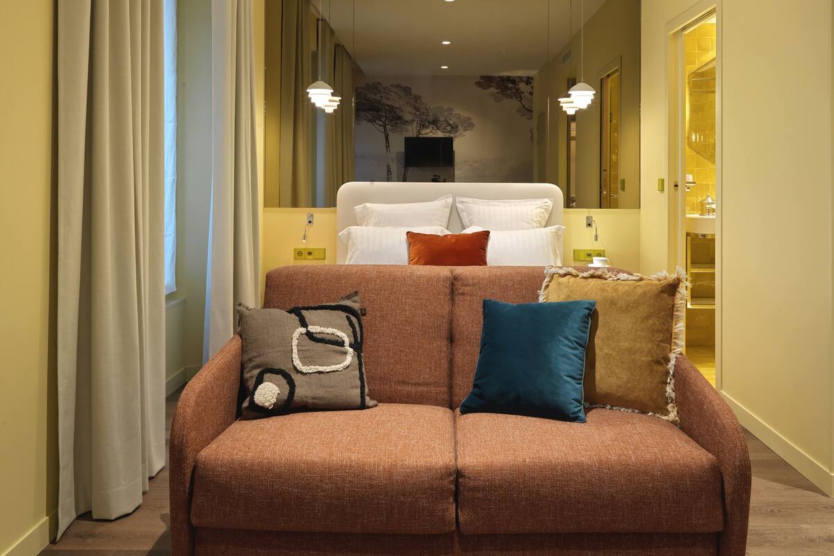 A modern hotel room with a brown sofa, a bed with white linens, and a large mirror.
