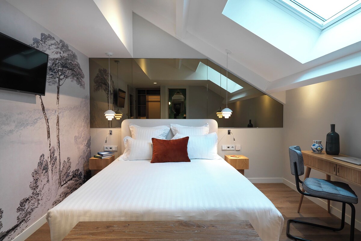 A modern bedroom with a skylight, a bed with white linens, a wooden nightstand, a desk with a chair, and a wall-mounted TV.