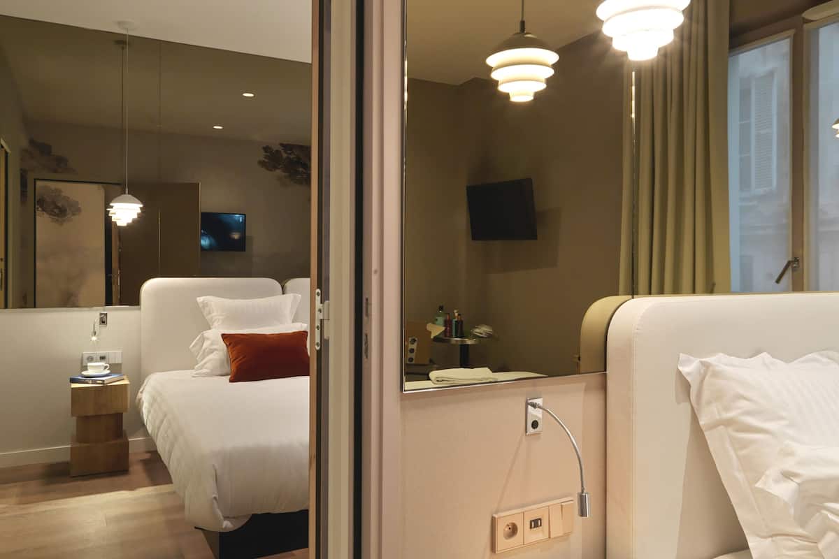 A hotel room with a bed, bedside table, and a mirror. There is a TV mounted on the wall and a window with curtains.