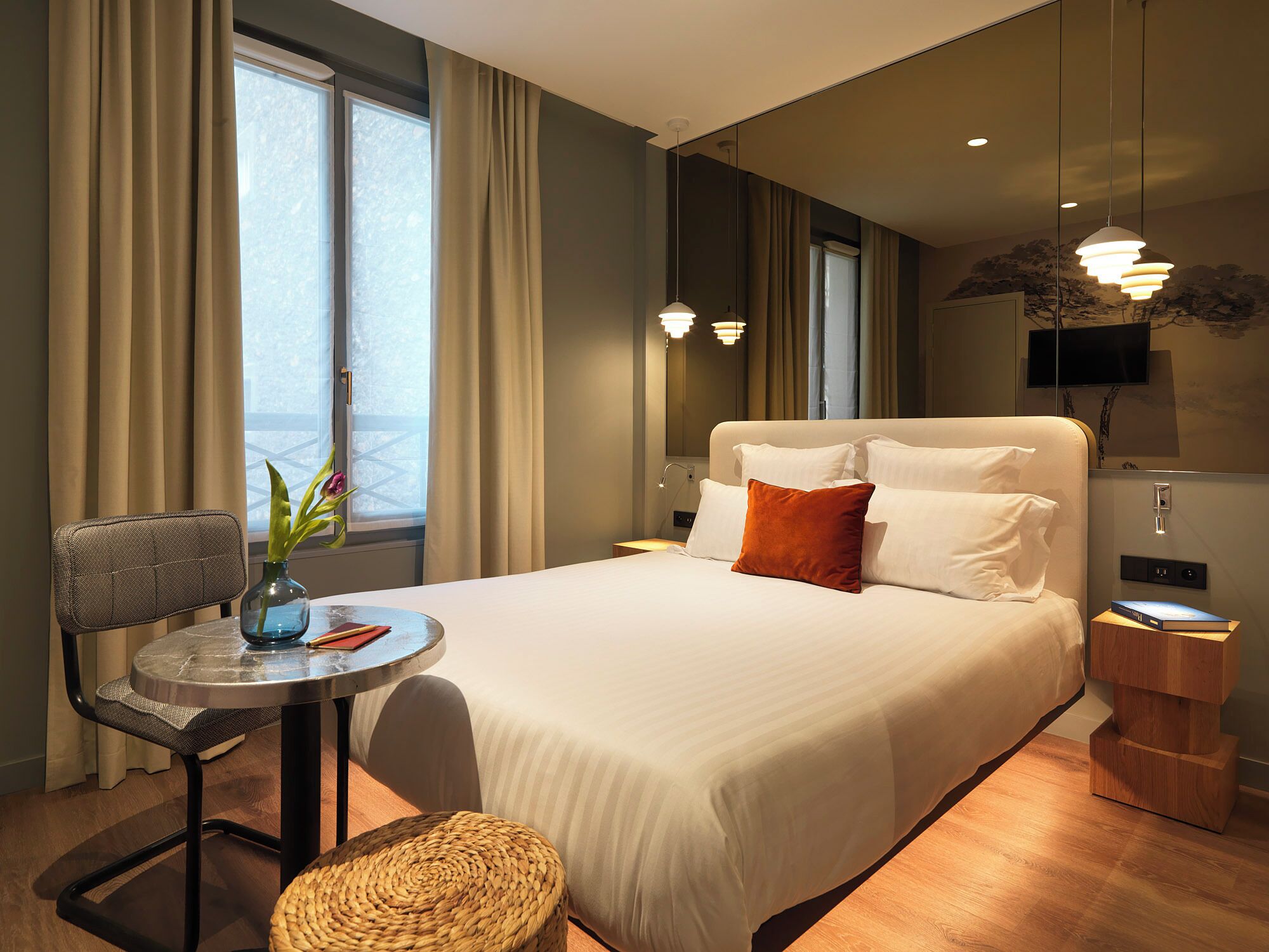 A hotel room with a bed, a small table with a chair, a TV, and a window with curtains.