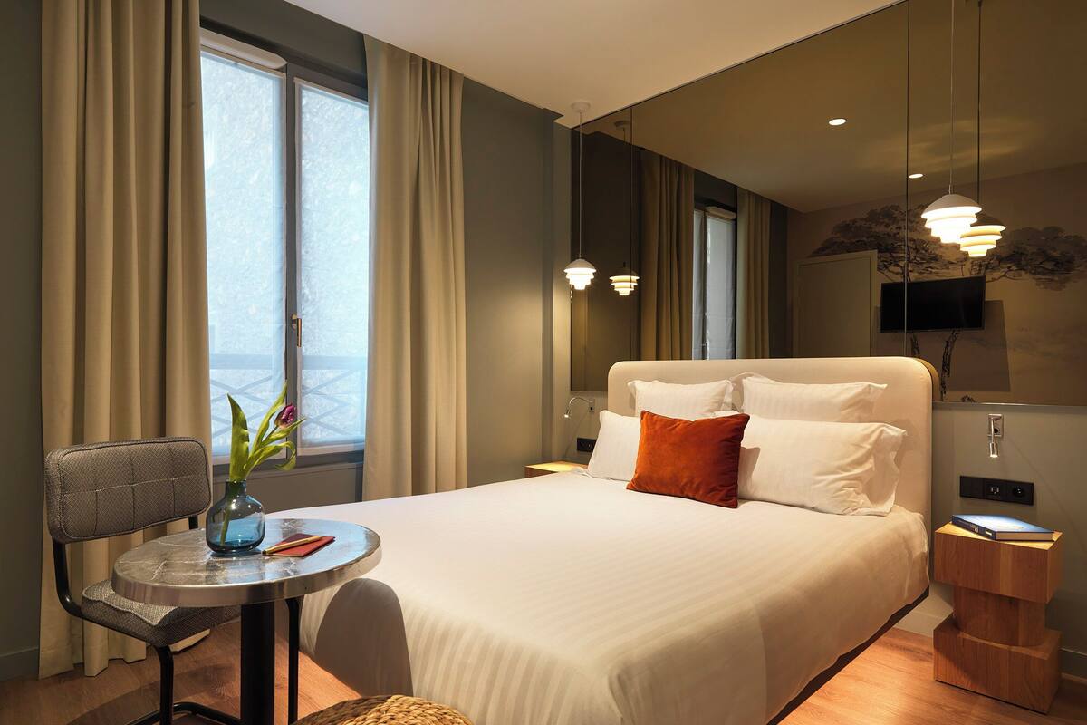 A hotel room with a bed, a small table with a chair, a TV, and a window with curtains.