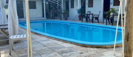 A pool area with a wooden swing, stone flooring, and a two-story building with balconies in the background.
