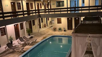 Une piscine dotée de chaises longues et d’un espace de repos couvert, le tout entouré d’un bâtiment comportant de nombreux balcons et portes.