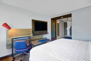 A modern hotel room with a large bed, a desk with a chair, a flat-screen TV mounted on the wall, and a shower area. - Tru by Hilton Stoughton (Stoughton)