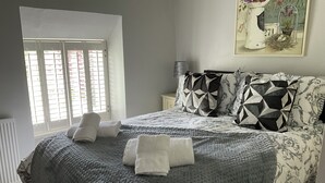 A bedroom with a large bed, two pillows, a bedside table, and a window with blinds. - Cosy luxurious 18th century stone built character cottage with stunning views (Penrith)