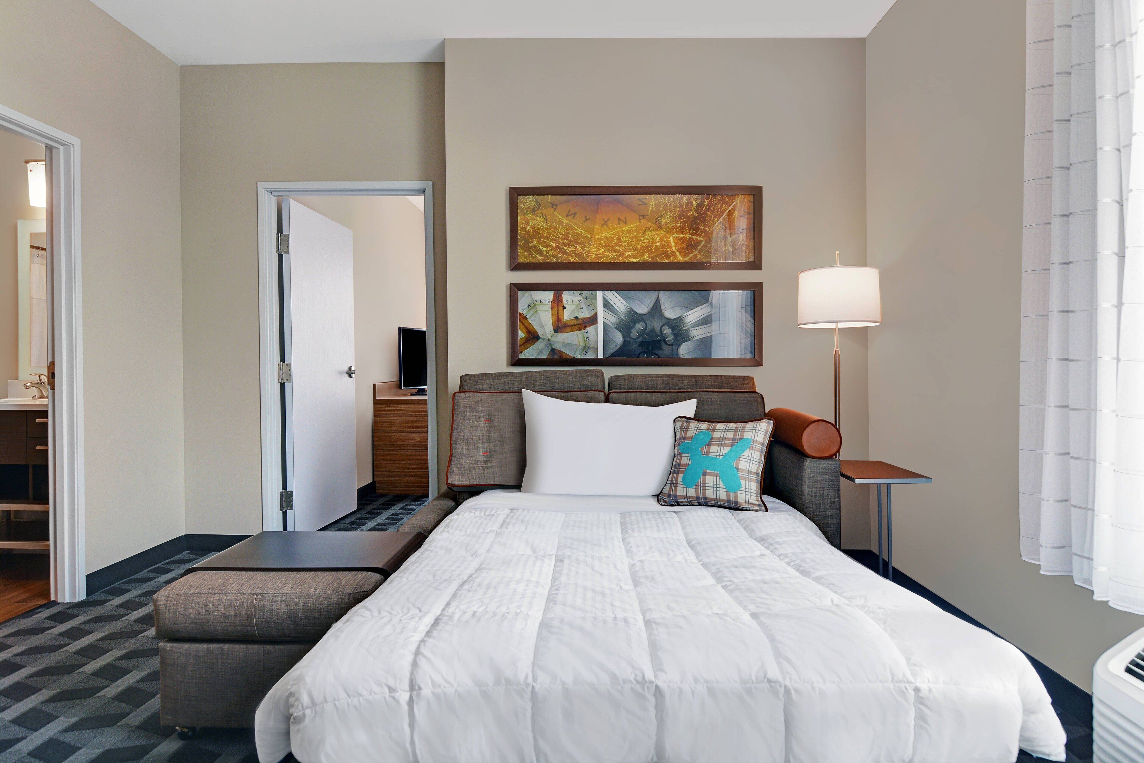 A hotel room with a large bed, a bedside table, a lamp, and two framed artworks on the wall.