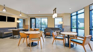 Een ruime binnenruimte met diverse zithoekopstellingen, waaronder ronde tafels en stoelen, een bank en een lange zitbank. Grote ramen zorgen voor natuurlijk licht en uitzicht op het omliggende landschap.