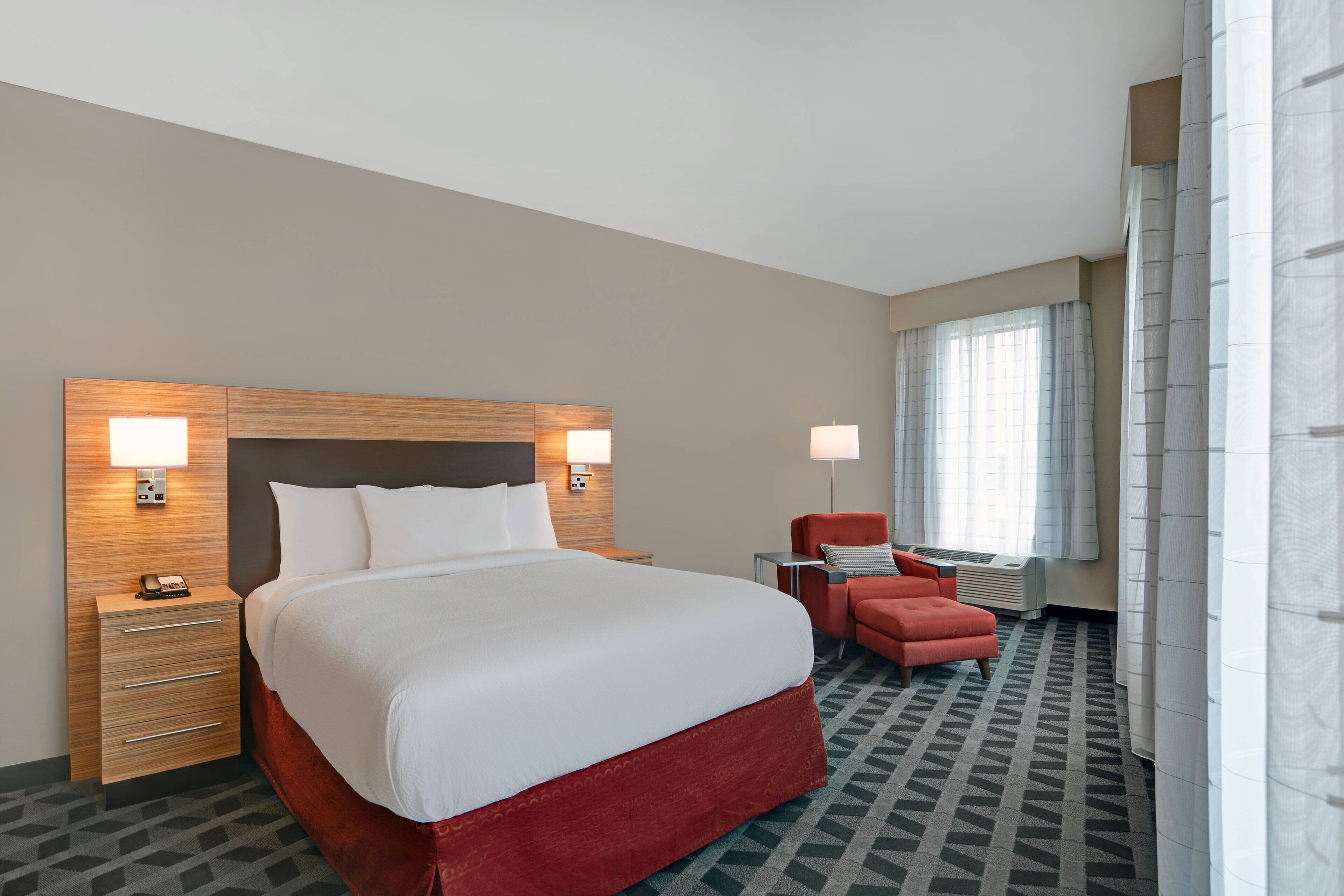 A hotel room with a large bed, a nightstand, a desk, a chair, and a window with curtains.