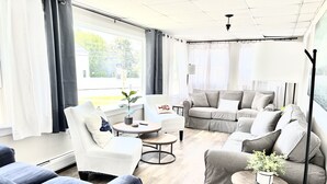 Ett modernt vardagsrum med en grå soffgrupp, en vit fåtölj och en blå fåtölj. Det finns ett litet runt bord med en växt och en lampa. Rummet har stora fönster med gardiner och utsikt över omgivningen.