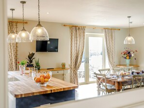 A dining area with a wooden table, chairs, a TV mounted on the wall, and a large window with curtains. - 4 bedroom accommodation in Draycott, near Cheddar (Draycott, near Cheddar)