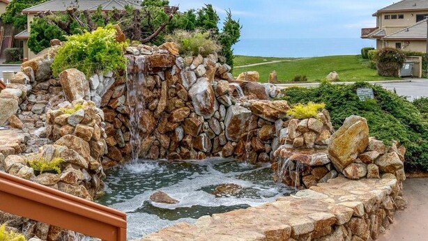 Une cascade artificielle en pierre, agrémentée d’un mur de pierres apparentes, aménagée dans un espace paysager verdoyant et offrant une vue sur un quartier résidentiel.