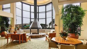 Un espace intérieur vaste et lumineux, avec un aménagement central autour d’un coin salon, de grandes fenêtres et des plantes en pot.