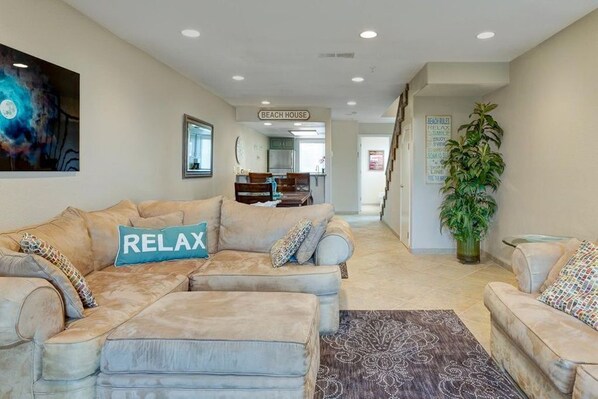 A living room with a beige sectional sofa, a 'RELAX' pillow, a large abstract painting, a 'BEACH HOUSE' sign, and a 'BEACH RULES' poster.