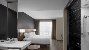 A modern hotel room with a large bed, a bedside table, a mirror, a bathroom with a shower, and a view of the city through the window. - AC Hotel Pittsburgh Southpointe (Canonsburg)