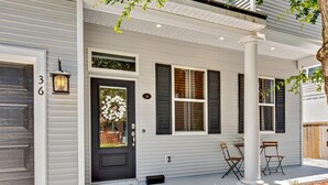 Una casa con puerta principal oscura, revestimiento blanco y contraventanas negras. Hay dos sillas y una pequeña mesa en el porche.